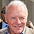 An older man with short white hair smiles while standing outside, wearing a light-colored jacket. A person in sunglasses is visible in the blurred background.